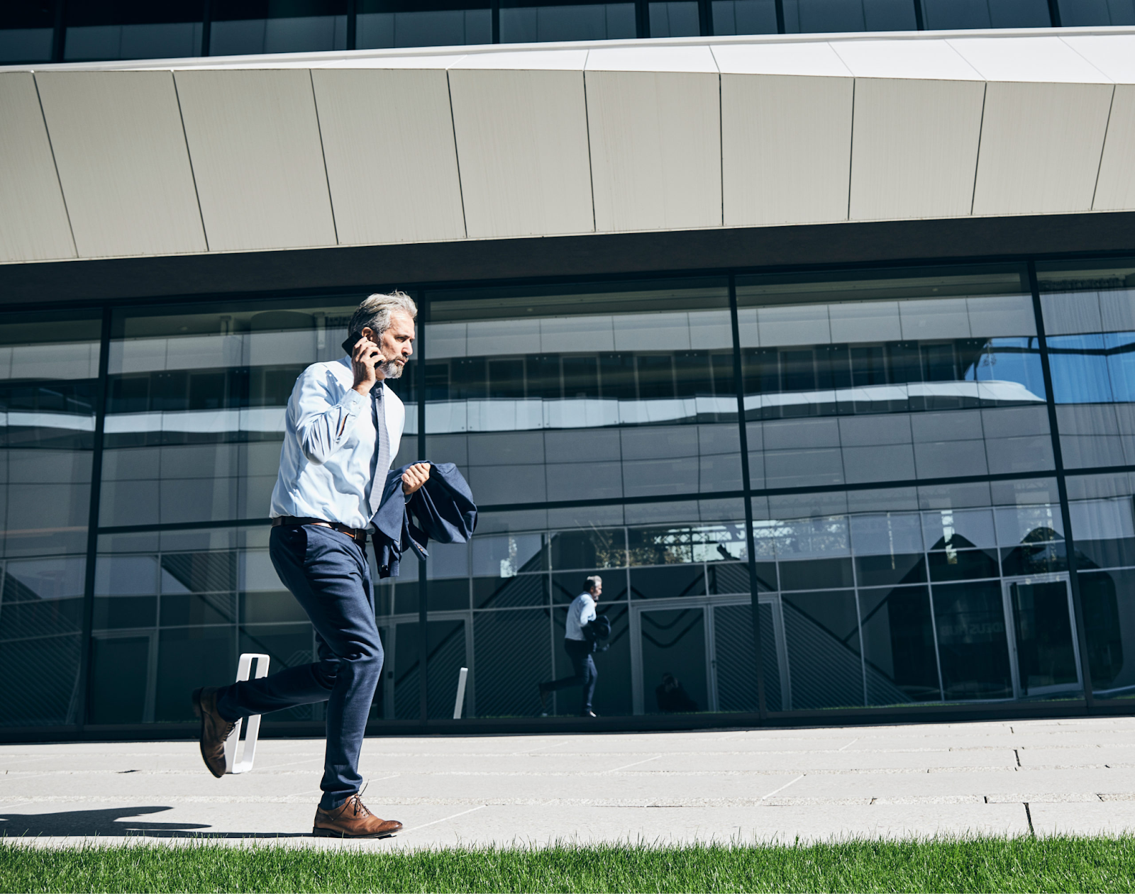 A man in a light blue shirt and dark pants walks briskly while talking on his phone, with a modern building in the ba…